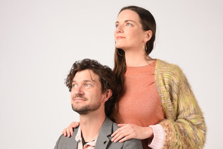 Indie pop duo Heaps Good Friends pose against a plain background, with a woman in a cardigan resting her hands on a man's shoulders as both look up.